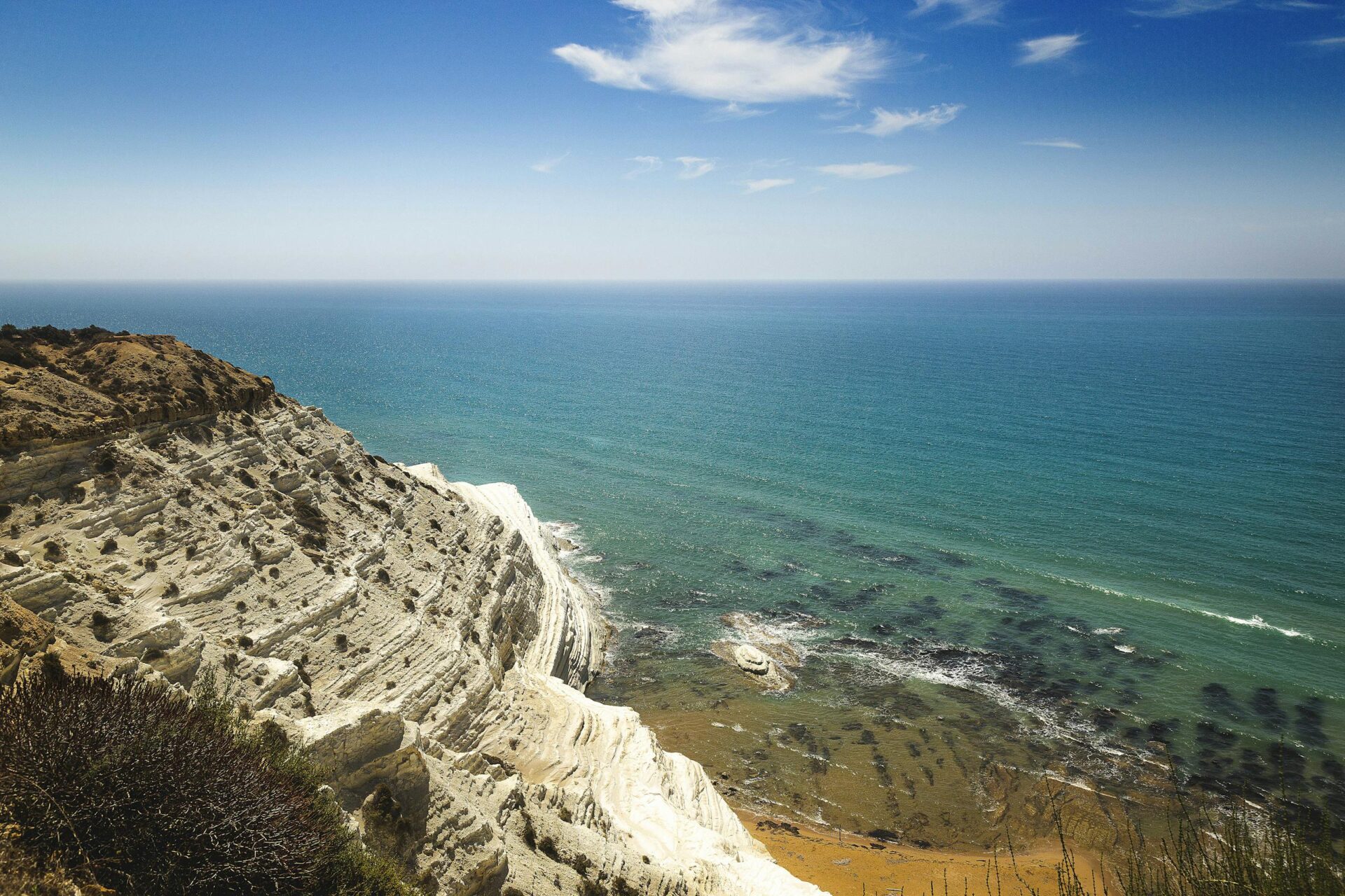 Stunning aerial view of a rocky coastal cliff with clear blue ocean under a bright sky.